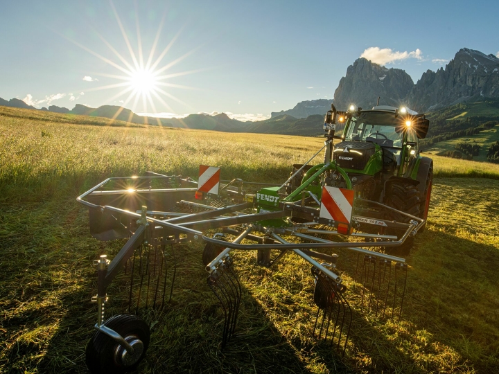 The green Fendt Former Alpine rake stands in front of a green Fendt tractor in a mountain meadow. In the background, the sun is slowly setting, the sky is blue and the mountains can also be seen