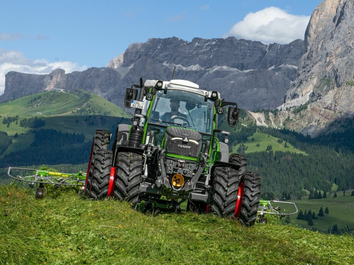 The Fendt 200 Vario with double tyres and red rims driving on a green mountain meadow, with mountains and blue sky in the background