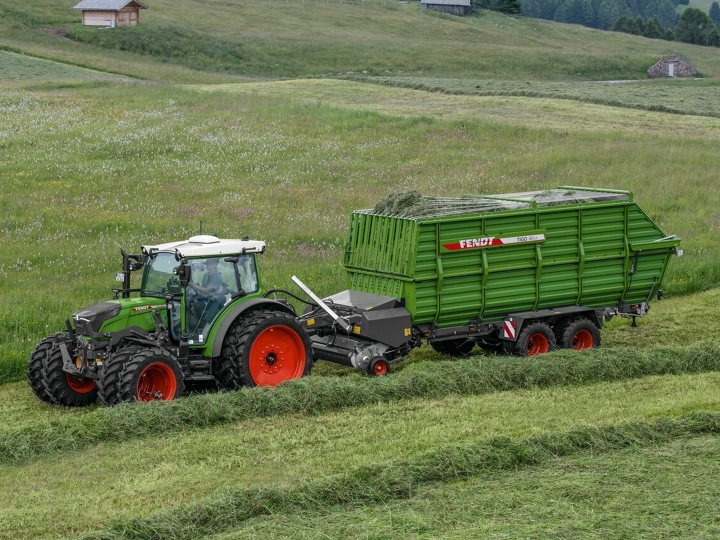 A green Fendt tractor with red rims and a green Fendt Tigo forage wagon with red rims driving on a green mountain meadow