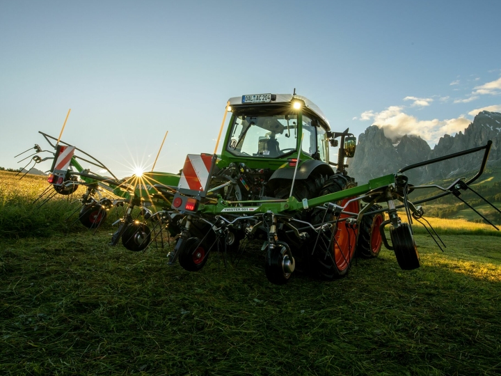 The green Fendt Twister hay tedder standing in front of a green Fendt tractor in a mountain meadow. In the background, the sun is slowly setting, the sky is blue and the mountains can also be seen