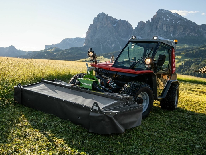 The Fendt Slicer Alpine mower in action on a mountain meadow with mountains and blue sky in the background