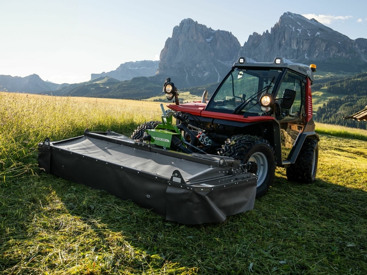 The Fendt Slicer Alpine mower in action on a mountain meadow with mountains and blue sky in the background