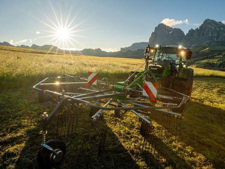 The green Fendt Former Alpine rake stands in front of a green Fendt tractor in a mountain meadow. In the background, the sun is slowly setting, the sky is blue and the mountains can also be seen