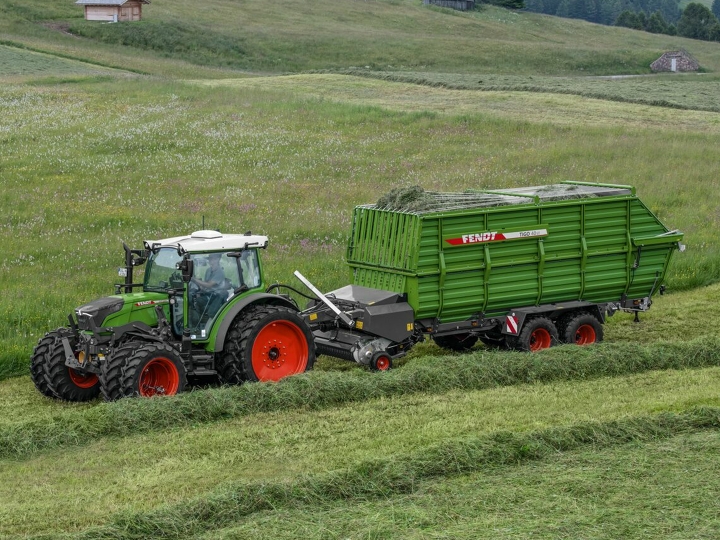 A green Fendt tractor with red rims and a green Fendt Tigo forage wagon with red rims driving on a green mountain meadow