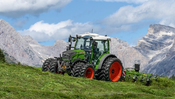 The green Fendt 200 Vario tractor with red rims and dual tyres is at work on a mountain meadow with a blue sky, white clouds and mountains in the background.