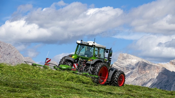 The Fendt Alpine tedder and the green Fendt tractor with red rims in action on a mountain meadow with a blue sky, white clouds and mountains in the background.