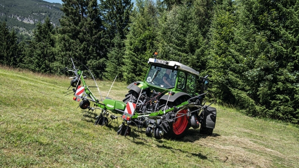 The folded Fendt Twister hay wagon is attached to a green Fendt tractor with red rims and driving across a meadow with a forest in the background