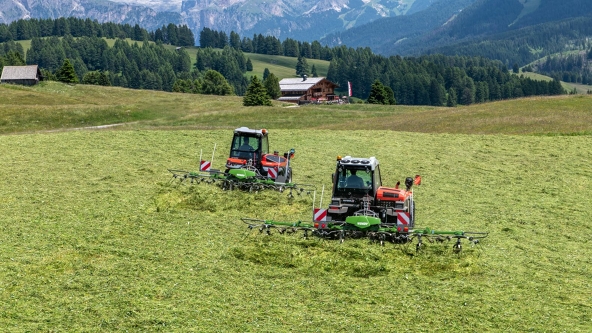 Two Fendt Twister Alpine tedders in action on a green mountain meadow. In the background are two huts and a forest