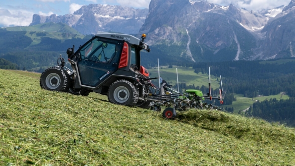 The green Fendt Twister tedding hay in a mountain meadow with a blue sky and mountain in the background.