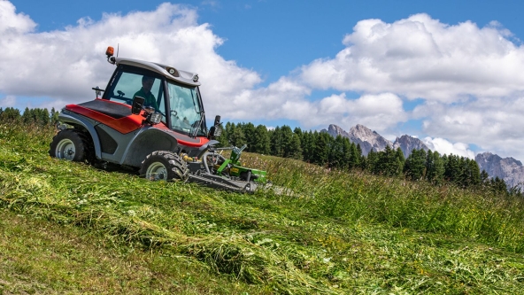 A green Fendt Slicer Alpine mowing a green meadow downhill, leaving a trail of freshly cut grass.