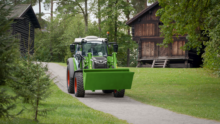 A Fendt e107 Vario driving down a road in Norway with a Fendt frontloader and bucket, wooden houses in the background