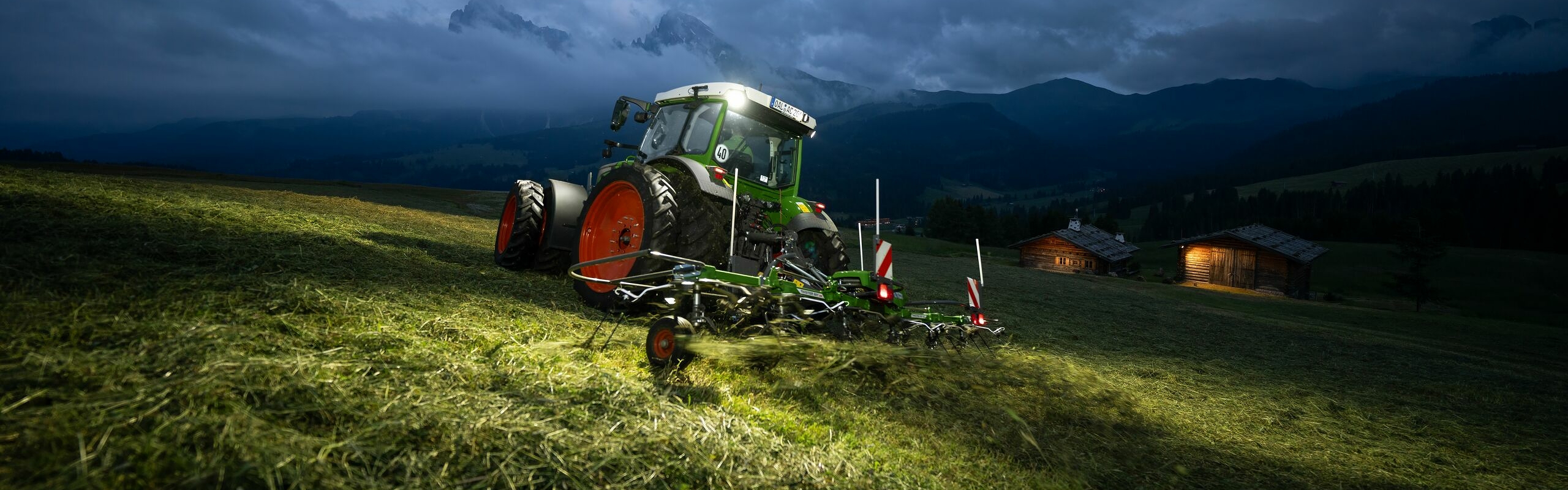 The green Fendt tractor with red rims and the green Fendt Twister implement travelling at an angle in a meadow in the mountains. Clouds, mountains and an illuminated hut can be seen in the background.