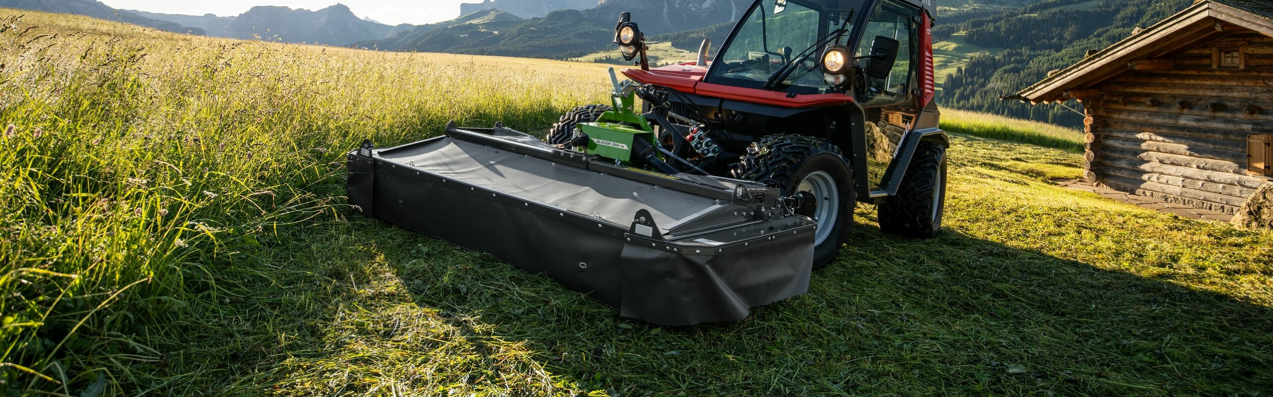 La falciatrice a dischi Fendt Slicer 260 FK verde durante l’utilizzo su un prato con monti, alberi e una baita marrone sullo sfondo.