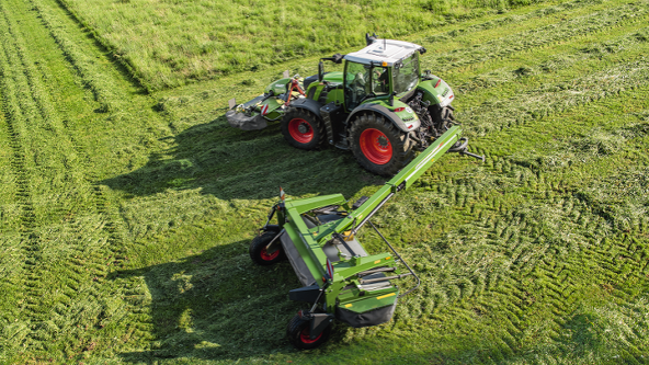 Un trattore Fendt verde con Fendt Slicer trainata con carrello di trasporto su una strada di campagna. Sullo sfondo ci sono il cielo blu con nuvole bianche e un bosco.