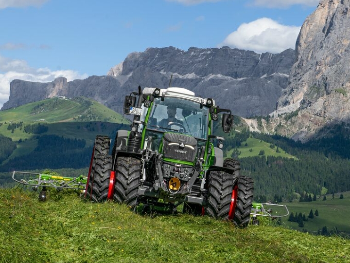 Il Fendt 200 Vario con pneumatici doppi e cerchi rossi viene utilizzato e procede su un prato di montagna verde, con monti e cielo azzurro sullo sfondo