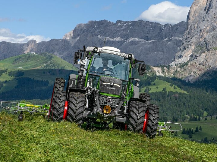 Il trattore Fendt 200 Vario verde con cerchi rossi e doppi pneumatici è al lavoro su un prato di montagna con un cielo blu, nuvole bianche e montagne sullo sfondo.