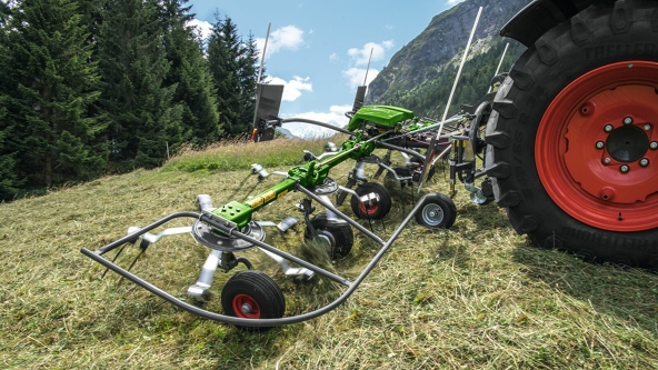 Due spandi-voltafieno Fendt Twister gamma Alpina durante l’uso su un prato di montagna verde. Sullo sfondo si trovano due baite e una foresta