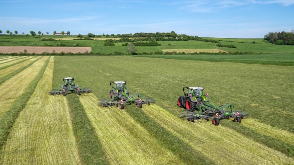 Trys "Fendt" traktoriai vienas šalia kito šienauja pievą, kiekvienas su "Fendt Former" grėbliu.