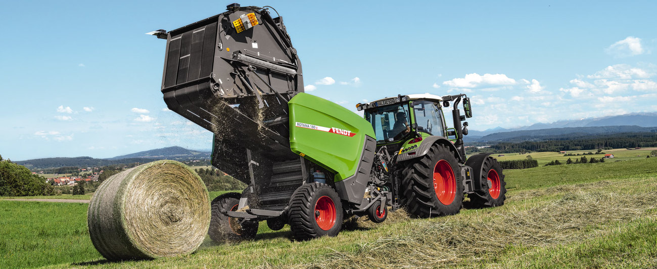 Fendt Traktor pļavā, presējot rulonus ar Fendt Rotana 180 V. Fonā redzami kalni.