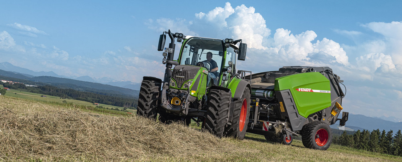 Fendt Rotana 180 V pļavā. Fona redzami kalni un debesis ar mākoņiem.