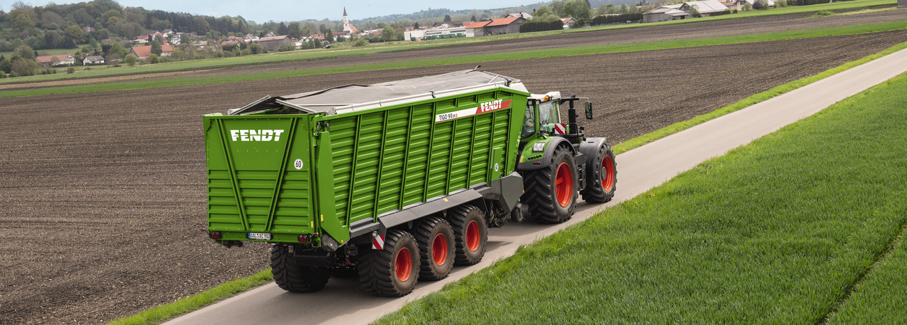 Fendt 900 Vario ar Fendt Tigo pašiekrāvējpiekabi aizmugures skats, transportējot zāli pa asfaltētu lauku ceļu.