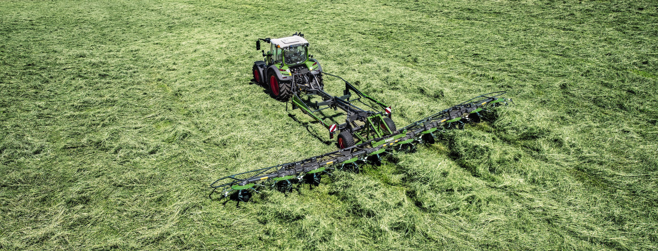 Zaļas krāsas Fendt traktors ar sarkaniem riteņu lokiem un zaļas krāsas Fendt Lotus agregāts darbībā zaļā pļavā zāles pļaušanas laikā