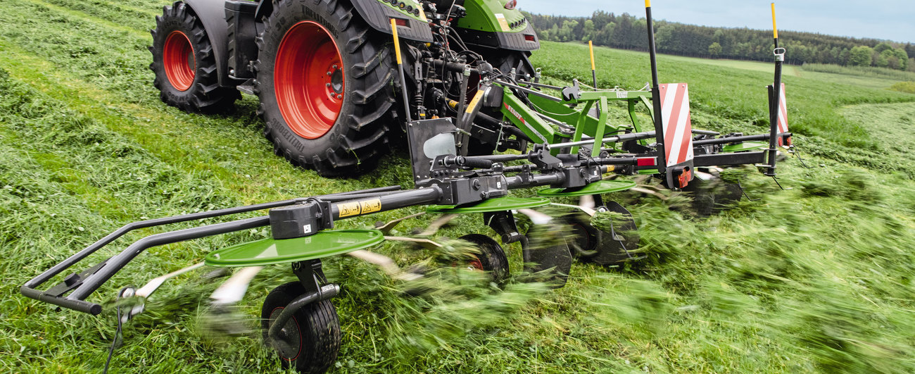 Jauns attēls ar zaļas krāsas Fendt Lotus agregātu un zaļas krāsas Fendt traktoru ar sarkanas krāsas riteņu lokiem, apgrozot zāli zaļā pļavā. Fonā skats uz zaļu ainavu un zilām debesīm.