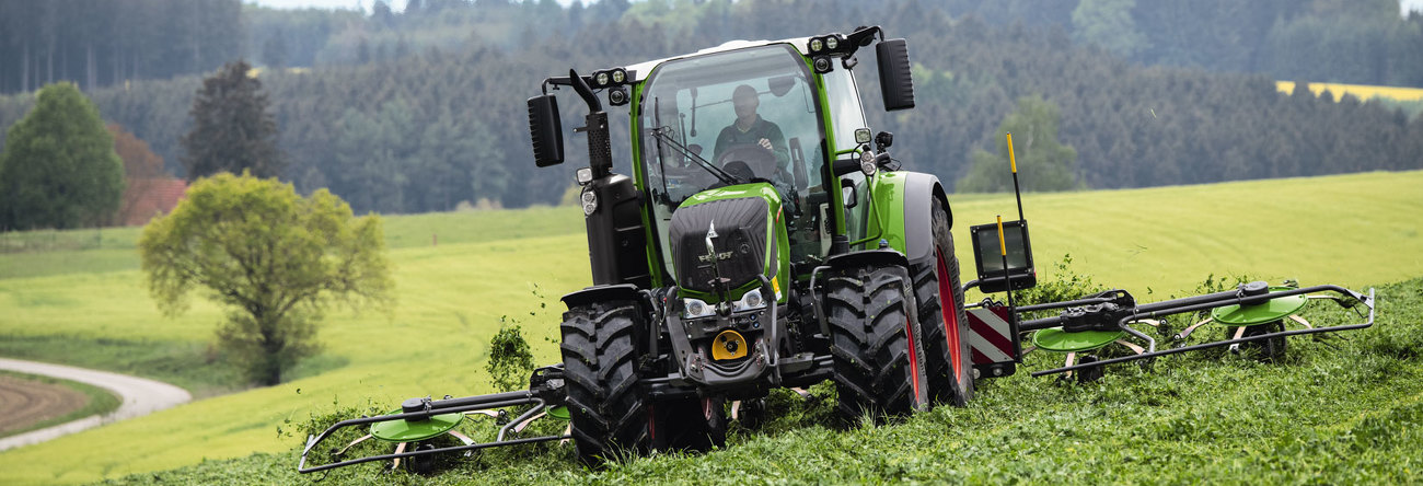 Zaļas krāsas Fendt traktors ar sarkaniem riteņu lokiem un zaļas krāsas Fendt Lotus agregāts zaļā pļavā zāles pļaušanas laikā un plašu lauku ainavu.