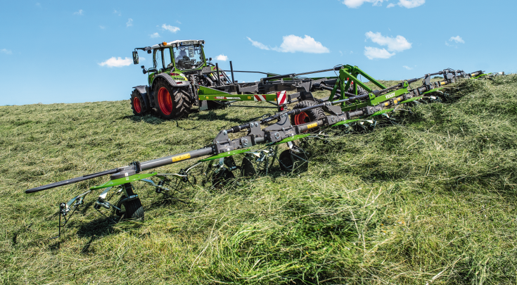 Zaļas krāsas Fendt traktors ar zaļas krāsas Fendt Lotus darbībā uz lauka, ārdot nopļauto zāli, ar zilām debesīm fonā.