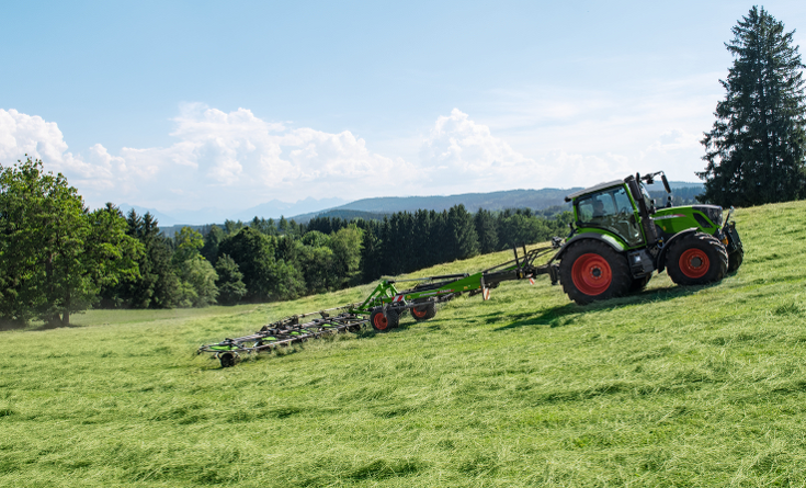 Zaļas krāsas Fendt traktors ar sarkaniem riteņu lokiem un zaļas krāsas Fendt Lotus agregāts zaļā pļavā zāles pļaušanas laikā ar zilām debesīm un plašu lauku ainavu.