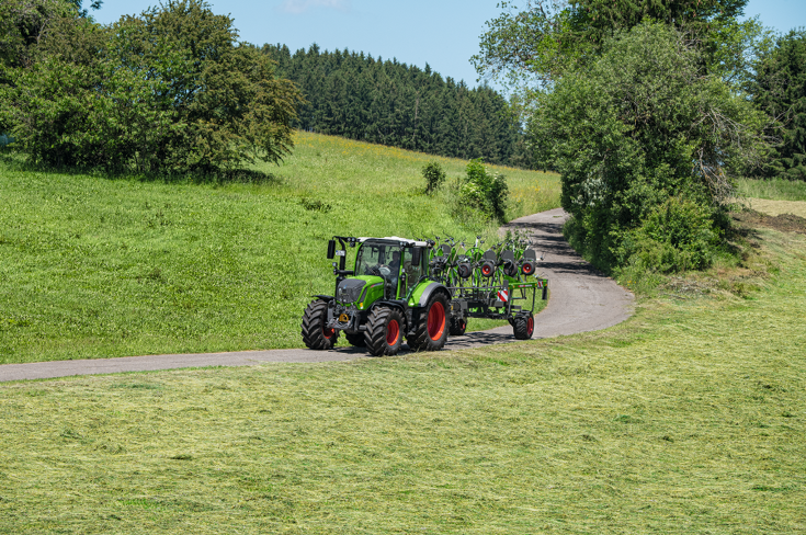 Zaļas krāsas Fendt traktors ar sarkaniem riteņu lokiem un zaļas krāsas Fendt Lotus agregāts brauc pa šauru celiņu zaļā pļavā, ar zilām debesīm un plašu lauku ainavu.