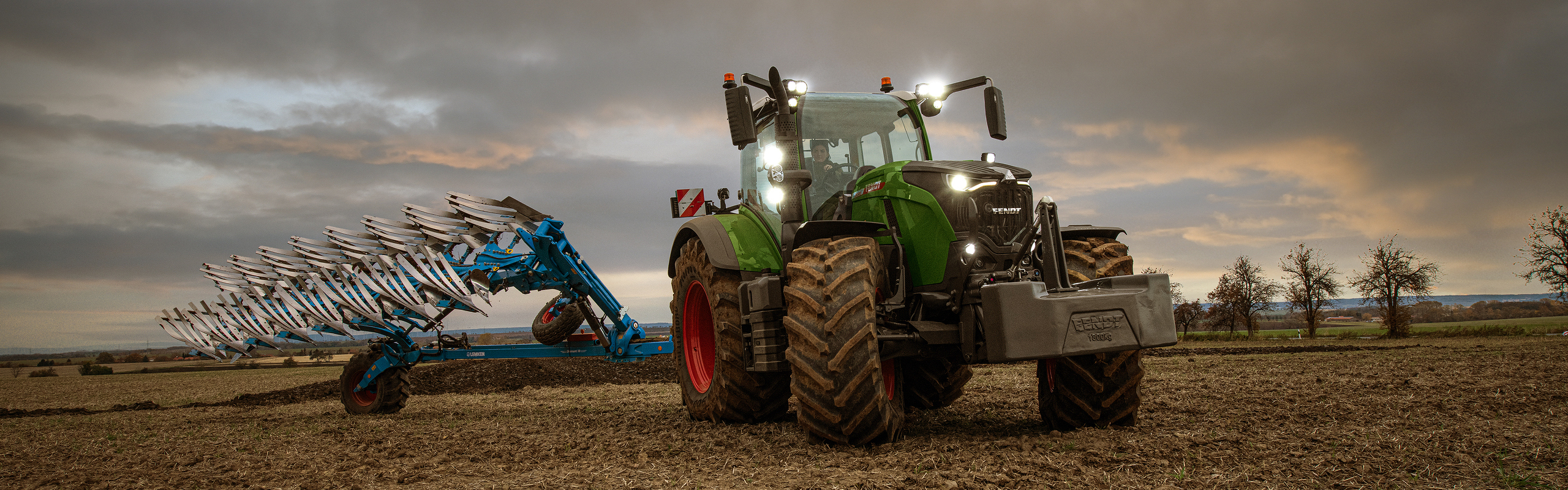 lauksaimniece brauc ar savu Fendt 700 Vario Gen7 uz lauka krēslas laikā.