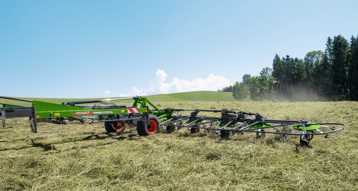 Zaļas krāsas Fendt Lotus siena ārdītājs darbībā zaļā pļavā, apgrozot sienu.