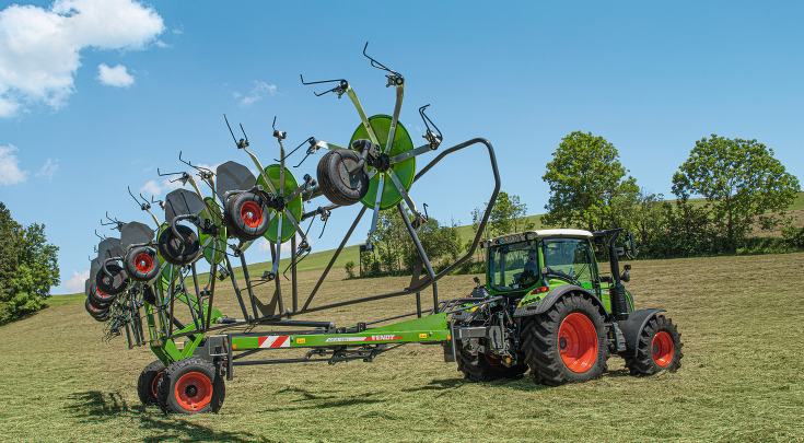 Zaļas krāsas Fendt traktors ar sarkaniem riteņu lokiem un zaļas krāsas Fendt Lotus salocītā stāvoklī zaļā pļavā ar zilām debesīm un skatu uz zaļu pakalnu.