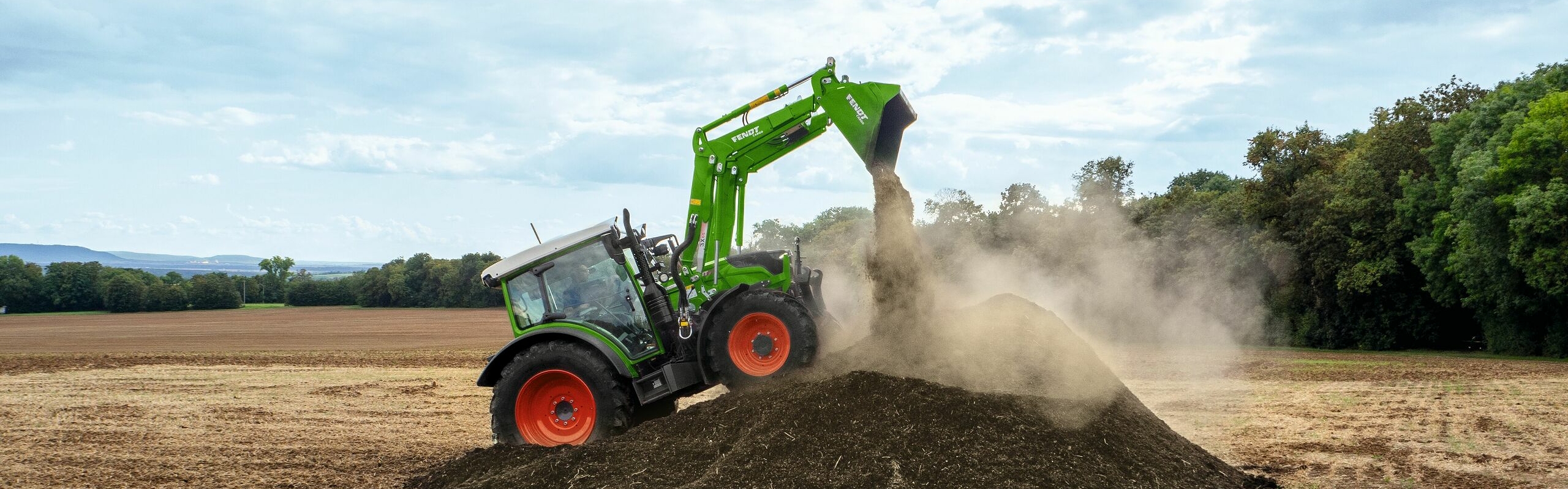 Fendt 200 Vario tīrumā ar Fendt Cargo, irdinot augsni. Priekšplānā redzami putekļi un fonā koki.