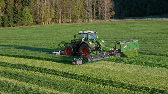 Zaļš Fendt Vario traktors ar sarkaniem atlokiem darba laikā pļavā ar uzstādītiem pļaušanas agregātiem.