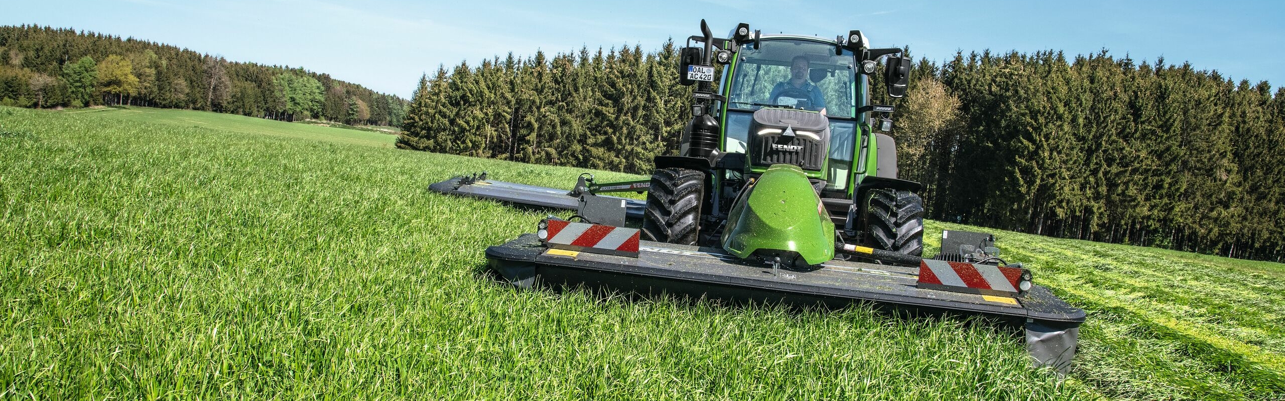 Fendt 200 Vario novāc zaļo lopbarību ar F sērijas priekšā uzstādamo pļaujmašīnu ar svārstīgo uzkari un aizmugurējo uzkari; fonā redzams mežs un zilas debesis.