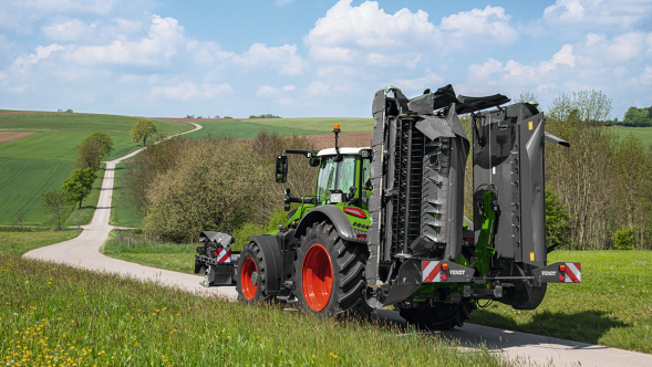 Zaļš Fendt Vario traktors uz lauka ceļa ar pļaujmašīnām transportēšanas pozīcijā.