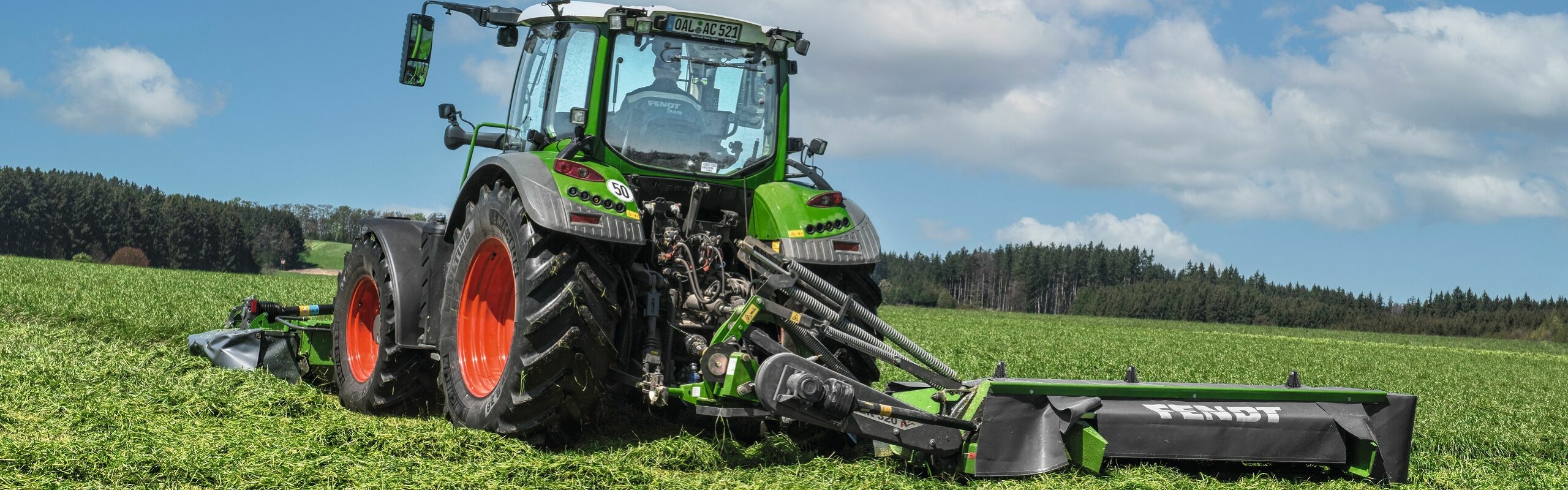 Zaļas krāsas Fendt traktors ar sarkaniem riteņu lokiem un Fendt Slicer P aizmugurējo uzkari zālāju platībā lopbarības novākšanas laikā.