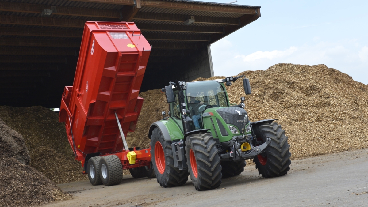 Fendt 700 Vario Gen5 traktors ar pašizgāzēju piekabi, kas noliktavā izkrauj šķeldas