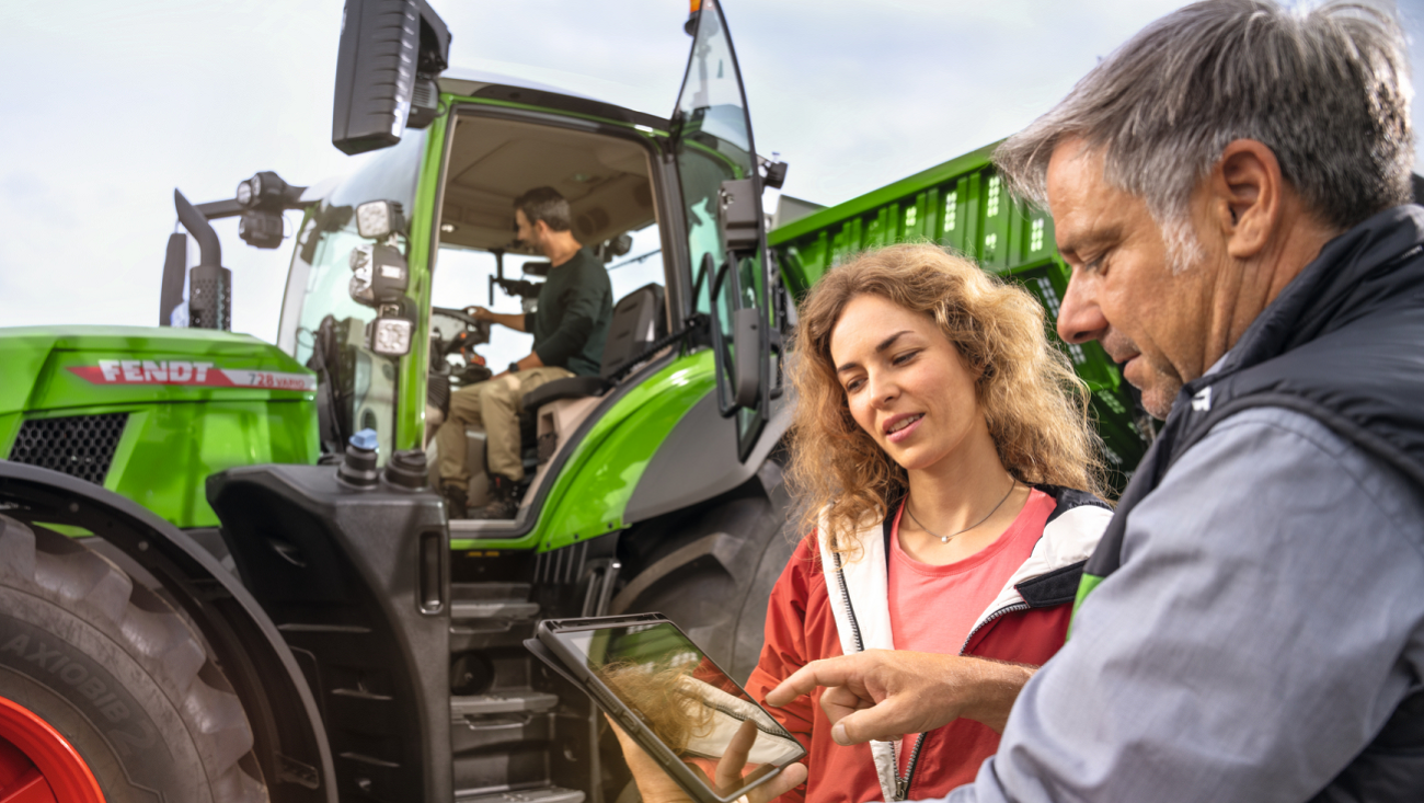 lauksaimnieks sēž Fendt 700 Vario Gen7, kamēr traktora priekšā viņa sieva konsultējas ar izplatītāju, turot rokās planšetdatoru.