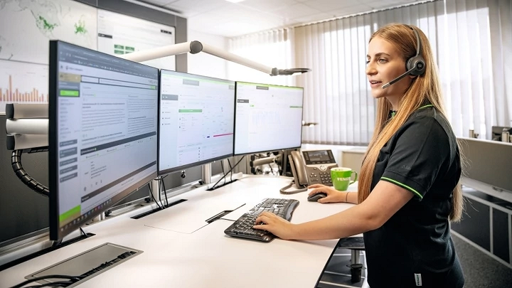 Een medewerkster van Fendt staat op kantoor met een headset op voor haar bureau en tuurt naar drie beeldschermen vol cijfers.