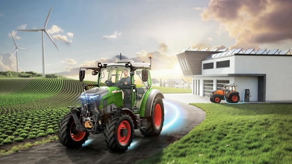 Een Fendt e100 Vario rijdend op een landweg nabij het dorp, terwijl op de achtergrond windmolens draaien en een andere Fendt e100 aan het opladen is