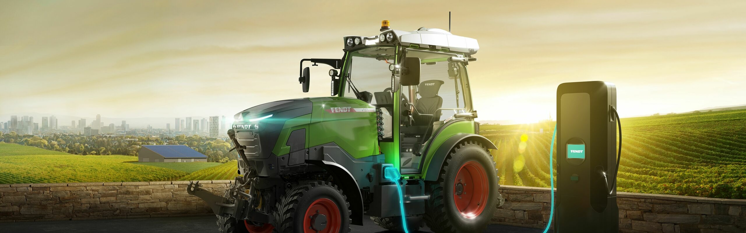 Een groene Fendt e100 V Vario met op de achtergrond weidegronden en daar weer achter het silhouet van de stad