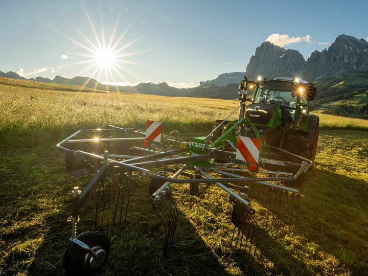 En grønn Fendt Former Alpine samlerive foran en grønn Fendt traktor på en fjelleng. I bakgrunnen går solen ned, himmelen er blå og fjellene kan også ses