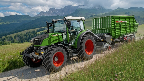 En grønnlakkert Fendt 200 Vario med Fendt Tigo lessevogn kjører på en fjellvei under fôrhøstingen.