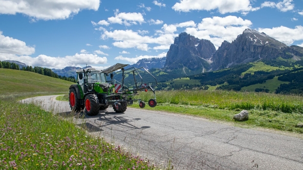 En grønn Fendt traktor med Fendt Former 351 DS i sammenlagt tilstand, kjører på en smal, grå vei. I bakgrunnen er det en grønn eng, fjellandskap og blå himmel med hvite skyer.