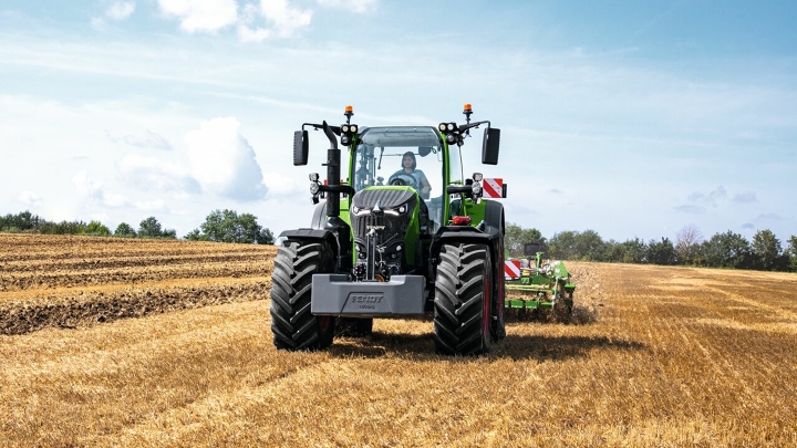 Ciągnik Fendt 700 Vario Gen 7 podczas pracy na polu. W tle drzewa i błękitne niebo z białymi chmurami.