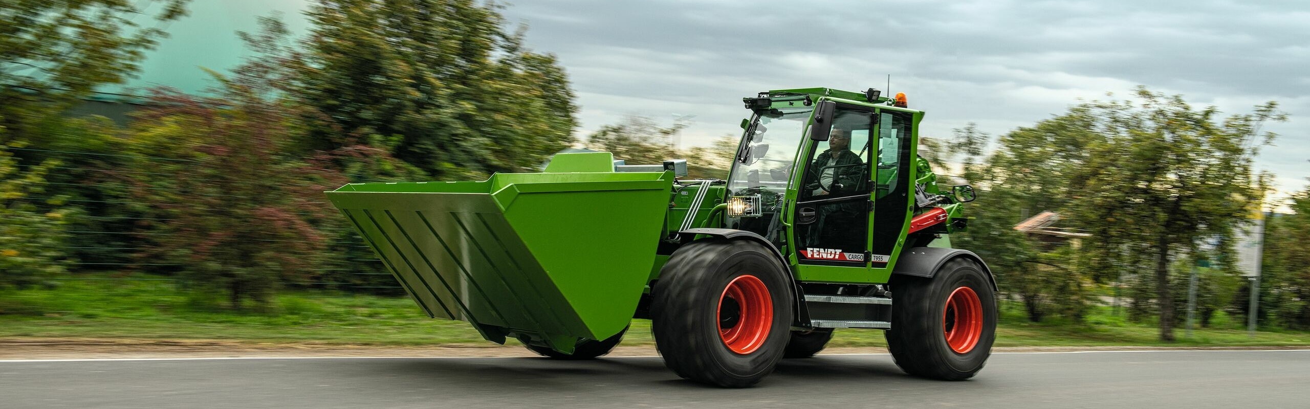 Rolnik prowadzi Fendt Cargo T955 po drodze, w tle widać lekko zamazane drzewa.