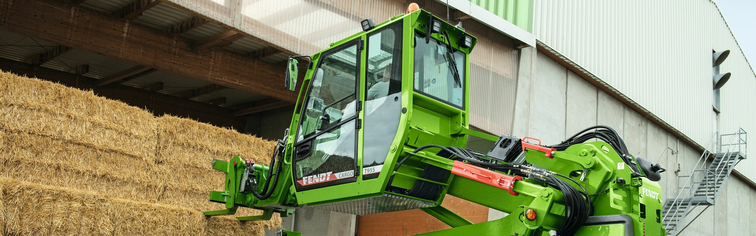 Zbliżenie z boku: rolnik siedzi w kabinie Fendt Cargo T955 i próbuje chwycić balot słomy łyżką.